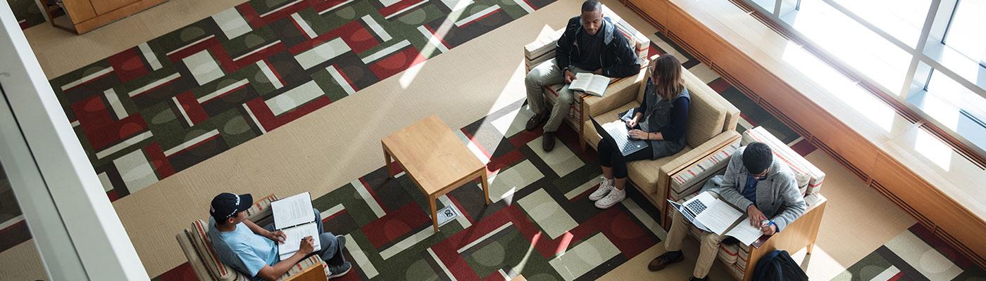 Overhead view of four students on comfy chars studying in the library lobby. Two of them are actively interacting.
